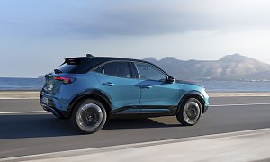 T&uuml;rkis-schwarzer Opel Mokka Electric f&auml;hrt an einer K&uuml;stenstra&szlig;e entlang, Meer und Gebirge im Hintergrund, Blick schr&auml;g von hinten links.