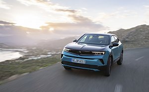 T&uuml;rkis-schwarzer Opel Mokka Electric f&auml;hrt bei Sonnenuntergang auf einer K&uuml;stenstra&szlig;e, Berge und Meer im Hintergrund, Blick frontal von vorn.