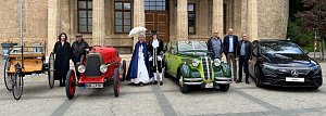 Gruppenfoto mit Fahrzeugen verschiedener Epochen: Vor einem historischen Gebäude stehen vier Autos – von einer Kutsche mit Holzaufbau über einen roten Oldtimer, einen grünen Oldtimer-Cabrio bis zu einem modernen schwarzen Mercedes. Davor posieren Personen in historischer Kleidung und moderner Garderobe.