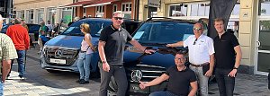Vier Männer posieren vor einem schwarzen Mercedes Vito auf einer Kopfsteinpflasterstraße in Heiligenstadt. Links und rechts im Hintergrund stehen weitere Fahrzeuge, im Hintergrund sind mehrstöckige Altstadthäuser mit Schaufenstern zu sehen.