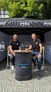 Infostand Autohaus Peter: Zwei M&auml;nner in schwarzen Polohemden mit Autohaus-Peter-Logo sitzen l&auml;chelnd an einem runden Stehtisch unter einem schwarzen Pavillon mit der Aufschrift Autohaus Peter Gruppe.