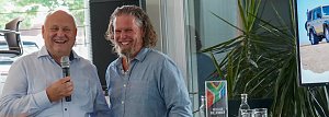 Afrikanischer Abend in Heiligenstadt: Helmut Peter und Abenteuer-Guide Hannu lächeln gemeinsam in die Kamera. Helmut Peter hält ein Mikrofon, im Hintergrund sind ein Bildschirm mit Geländewagen und eine Pflanze zu sehen.