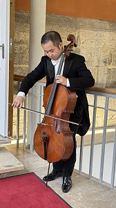 Ein Musiker in einem schwarzen Frack spielt konzentriert auf einem Cello. Die Szene spielt im Innenbereich eines historischen Geb&auml;udes mit Steinw&auml;nden und einem rotbraunen Teppich. Der Musiker steht am Gel&auml;nder einer Galerie, w&auml;hrend er mit dem Bogen &uuml;ber die Saiten des Instruments streicht.
