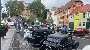 Blick auf eine Oldtimerschau in der Altstadt von Sondershausen. Mehrere historische Fahrzeuge sind auf dem Kopfsteinpflaster ausgestellt. Im Vordergrund klassische Mercedes- und Opel-Modelle, daneben Beachflags mit den Logos von Mercedes-Benz, Opel und Autohaus Peter. Im Hintergrund Fachwerkh&auml;user und das Schloss Sondershausen. Besucher schlendern durch die Ausstellung.
