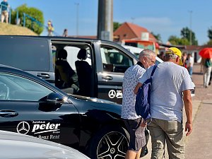 Zwei &auml;ltere M&auml;nner gehen nebeneinander &uuml;ber das Veranstaltungsgel&auml;nde des Leichtathletik-Meetings Anhalt 2025, vorbei an schwarzen Mercedes-Benz Fahrzeugen vom Autohaus Peter, darunter ein Van mit ge&ouml;ffneter Schiebet&uuml;r. Im Hintergrund sind weitere Besucher, Geb&auml;ude und ein sonniger, klarer Himmel zu sehen.