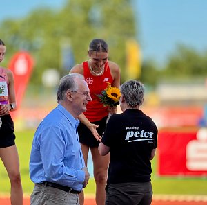 Bei der Siegerehrung des Anhalt 2025 Leichtathletik-Meetings &uuml;berreicht eine Mitarbeiterin des Autohauses Peter einer Athletin mit rotem Trikot und Sonnenblumenstrau&szlig; die Auszeichnung. Daneben steht ein &auml;lterer Mann im blauen Hemd, w&auml;hrend im Hintergrund die Laufbahn und farbige Werbebanner sichtbar sind.