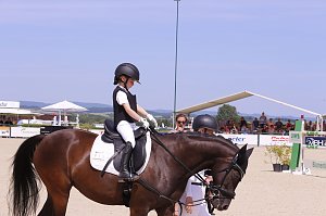 Ein Kind in wei&szlig;er Reitkleidung und schwarzer Weste sitzt auf einem braunen Pferd auf dem Turnierplatz in Immenrode. Vor dem Pferd f&uuml;hren zwei Erwachsene das Tier, im Hintergrund sind Zuschauertrib&uuml;nen und Hindernisse zu sehen.
