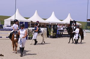 Mehrere Kinder reiten auf Ponys beim Reitturnier in Immenrode, begleitet von Erwachsenen in Reitkleidung. Im Hintergrund stehen wei&szlig;e Pagodenzelte und ein Hindernis mit Mercedes-Benz-Werbung.