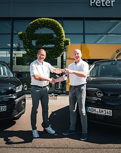 Vor dem Autohaus Peter stehen zwei M&auml;nner zwischen zwei schwarzen Opel-Fahrzeugen. Einer der M&auml;nner &uuml;berreicht dem anderen einen Autoschl&uuml;ssel, beide l&auml;cheln und geben sich die Hand. Im Hintergrund ist das Opel-Logo aus gr&uuml;nem Pflanzenmaterial an der Glasfassade zu sehen. Die Szene wirkt freundlich und sonnig.