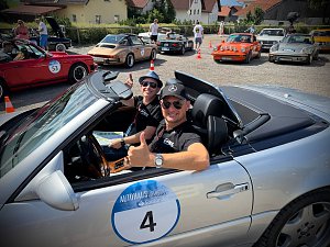Andreas Peter und Carsten Dost sitzen l&auml;chelnd in einem silbernen Mercedes-Benz SL 320 Cabrio mit Startnummer 4 bei einer Oldtimer-Rallye. Beide tragen Sonnenbrillen und zeigen Daumen nach oben. Im Hintergrund stehen mehrere weitere klassische Autos, darunter Porsche-Modelle, auf einem Parkplatz in einer sonnigen Dorfszene. Menschen unterhalten sich und bereiten sich auf die Veranstaltung vor.