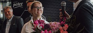 Lilly Hecker, frisch ausgebildete Automobilkauffrau und Jahrgangsbeste, steht lächelnd mit einem Blumenstrauß neben Geschäftsführer Andreas Peter, der in ein Mikrofon spricht. Im Hintergrund sind weitere Personen und das Logo der Autohaus-Peter-Gruppe zu sehen.