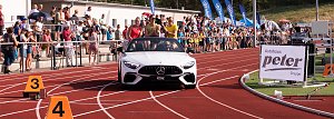 Weißes Mercedes-Cabrio fährt auf die rote Tartanbahn des Stadions ein. Auf dem Rücksitz sitzen mehrere Sportlerinnen, während Zuschauer am Rand applaudieren.