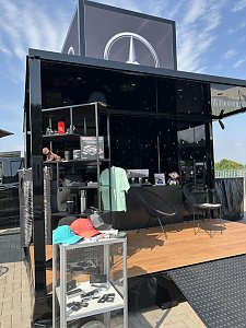 Schwarzer Mercedes-Stand mit gro&szlig;em Mercedes-Logo auf dem Dach. Im Vordergrund ein Glasdisplay mit Schl&uuml;sselanh&auml;ngern und Caps in Schwarz, Rot und T&uuml;rkis mit Mercedes-Stern. Dahinter Regale mit Merchandise-Artikeln wie Thermobecher, Modellautos, Kleidung und einem Stofftier. Zwei schwarze St&uuml;hle und ein Tisch stehen auf der h&ouml;lzernen Ausstellungsfl&auml;che.