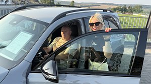 Ein Interessent sitzt am Steuer eines silbernen Mercedes GLB 220 4MATIC, w&auml;hrend ihn eine Peter-Mitarbeiterin mit Sonnenbrille und schwarzem Polohemd freundlich ber&auml;t. Auf der Windschutzscheibe ist ein Datenblatt mit Fahrzeugdetails sichtbar. Im Hintergrund ein Stadion mit Trib&uuml;ne und gr&uuml;ner Rasen..