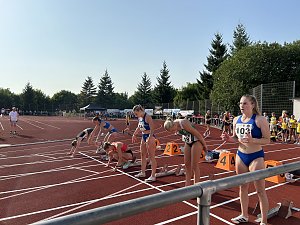 Mehrere Athletinnen bereiten sich auf der Tartanbahn an den Startbl&ouml;cken vor. Einige gehen bereits in die Hocke, andere stehen noch. Zuschauer im Hintergrund.