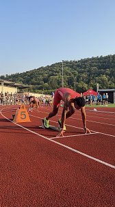 Sprinter im roten Trikot steht im Startblock auf Bahn 5, bereit f&uuml;r den Lauf. Weitere Athleten bereiten sich auf den benachbarten Bahnen vor. Zuschauer und Kampfrichter am Rand der Tartanbahn, im Hintergrund gr&uuml;ne H&uuml;gel.