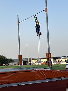 Stabhochspringer in blau-gelbem Trikot in der Luft kopf&uuml;ber &uuml;ber der Latte. Im Hintergrund das Stadion und Zuschauer.
