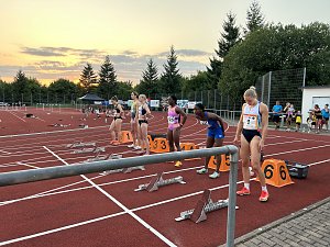 Sechs Sprinterinnen stehen an ihren Startbl&ouml;cken auf der Laufbahn. Der Himmel im Hintergrund zeigt einen Sonnenuntergang mit Wolken.