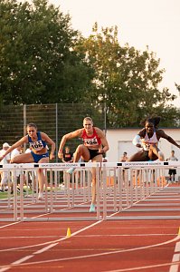 Drei Athletinnen im H&uuml;rdenlauf springen gleichzeitig &uuml;ber eine H&uuml;rde auf der roten Tartanbahn. Im Vordergrund sind die H&uuml;rden mit der Aufschrift �Sportzentrum am G&ouml;ldner� zu sehen. Zuschauer und Kampfrichter im Hintergrund.