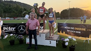 Drei Sportlerinnen stehen nach einem Leichtathletik-Wettkampf auf einem Siegerpodest und halten Blumenstr&auml;u&szlig;e in den H&auml;nden. Davor steht Centerleiter Marko Wuttke. Im Hintergrund sind eine gr&uuml;ne Sportanlage, Zuschauer sowie eine Waldkulisse bei Sonnenuntergang zu sehen. Vorne am Podest h&auml;ngen Werbebanner, unter anderem vom Autohaus Peter und Mercedes-Benz.