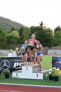 Drei Athletinnen stehen auf dem Siegerpodest, alle halten Blumenstr&auml;u&szlig;e. Die Siegerin in der Mitte tr&auml;gt ein rotes Trikot, die beiden anderen blaue Outfits. Im Hintergrund das Stadion und gr&uuml;ne H&uuml;gel.