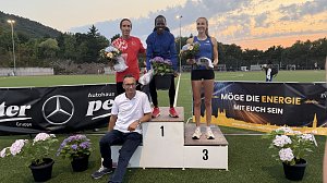 Drei Athlet:innen stehen auf dem Siegerpodest mit Blumen in den H&auml;nden, vor ihnen Mercedes-Benz Pkw Verkaufsleiter J&ouml;rn Bierwisch mit wei&szlig;em Shirt. Hinter dem Podest Sponsorenbanner und Abendhimmel.