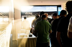 Eine Gruppe M&auml;nner steht in einer Schlange am Buffet. Sonnenlicht f&auml;llt durch die T&uuml;r, im Hintergrund ein Pavillon mit Autohaus-Peter-Logo.