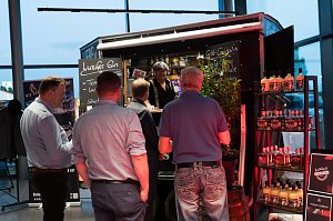 Mehrere M&auml;nner stehen an einem Gin-Stand mit schwarzem Verkaufswagen. Auf Schildern steht Luzifer Gin und Gin-Cocktails. Regale mit Flaschen sind daneben aufgestellt.
