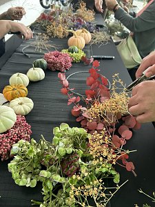 Auf einem Tisch liegen kleine K&uuml;rbisse, bunte Beeren und verschiedene Zweige f&uuml;r herbstliche Dekoration. Mehrere Personen gestalten daraus Kr&auml;nze, einige H&auml;nde sind bei der Arbeit zu sehen.
