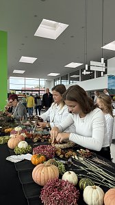 Besucherinnen gestalten an einem Basteltisch herbstliche Kr&auml;nze mit K&uuml;rbissen, Beeren und Zweigen. Auf dem Tisch liegen bunte Naturmaterialien.