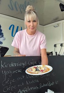 Frau in rosa T-Shirt pr&auml;sentiert einen frisch zubereiteten Cr&ecirc;pe mit Obst und Puderzucker. Im Hintergrund ist eine Tafel mit Cr&ecirc;pe-Sorten beschrieben.