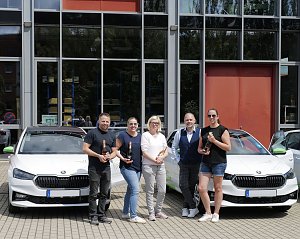 F&uuml;nf Personen stehen vor einem modernen Geb&auml;ude und posieren mit Champagnerflaschen vor zwei wei&szlig;en �koda-Fahrzeugen. Auf den Autos sind gr&uuml;ne Elemente und das Logo der Praxisklinik f&uuml;r Zahnheilkunde zu sehen.