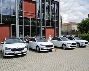 Vier wei&szlig;e �koda-Fahrzeuge mit gr&uuml;nen Akzenten stehen nebeneinander vor einem modernen Geb&auml;ude. Vor den Autos stehen mehrere Personen mit Flaschen in der Hand, wie bei einer feierlichen Fahrzeug&uuml;bergabe.
