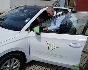 Ein Mann im blauen Sakko zeigt einer Frau auf dem Fahrersitz eines wei&szlig;en �koda-Fahrzeugs etwas auf einem Tablet. Auf der Autot&uuml;r ist das Logo der Praxisklinik f&uuml;r Zahnheilkunde zu sehen.