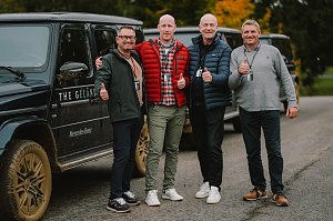 Vier M&auml;nner stehen l&auml;chelnd vor einem schwarzen Mercedes-Benz G-Class mit schlammigen Reifen. Alle tragen Freizeitkleidung und zeigen den Daumen nach oben. Hinter ihnen sind weitere Fahrzeuge und herbstlich gef&auml;rbte B&auml;ume zu sehen.