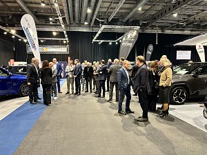 Viele Messebesucher stehen zwischen ausgestellten Autos und Markenflaggen und unterhalten sich in der Ausstellungshalle.