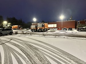 Winterliche Szene auf einem Betriebshof bei Nacht mit mehreren orangefarbenen Müllfahrzeugen und verschneitem Parkplatz.