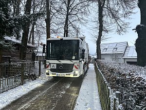 Müllfahrzeug fährt auf einer schmalen, verschneiten Straße zwischen Häusern und Bäumen.