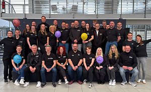 Man sieht ein Teamfoto mit vielen Menschen auf und vor einer kleinen Bühne. Alle haben das gleiche T-Shirt an, manche halten bunte Luftballons hoch.