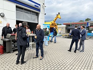 Hof vor Autohaus mit Grill, Hüpfburg im Hintergrund und einigen Personen, die sich unterhalten.