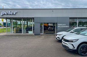 Der Eingangsbereich des Autohauses Peter in Halle Neustadt/ Göttinger Bogen mit großer Glasfront und der Aufschrift "Herzlich Willkommen". Rechts vor dem Gebäude sind zwei weiße Opel-Modelle zu sehen.