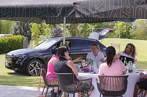 Gäste sitzen unter Sonnenschirmen zusammen und genießen Gespräche auf der Terrasse neben einem Mercedes SUV.