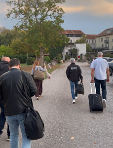 Mehrere Personen gehen von hinten zu sehen mit Gepäck einen asphaltierten Weg entlang. Links läuft eine Frau mit Schultertasche, rechts ein Mann in schwarzer Jacke mit Peter-Schriftzug sowie ein Mann, der einen Rollkoffer zieht. Im Hintergrund steh