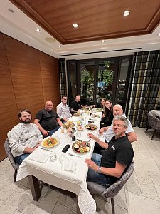 Acht Personen sitzen gemeinsam an einem Tisch in einem eleganten Restaurant mit Holzvertäfelung und Blick ins Grüne. Vor ihnen stehen Teller mit warmen Speisen. Die Gruppe blickt lächelnd direkt in die Kamera.