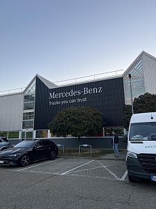 Ein modernes Gebäude mit einer dunklen Fassade trägt den großen weißen Schriftzug Mercedes-Benz Trucks you can trust. Davor erstreckt sich ein gepflasterter Parkplatz.