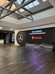 Eine große, beleuchtete Medienwand steht in einer modernen Halle mit einer verglasten Deckenkonstruktion und dunklem Holzboden. Auf dem Bildschirm prangt das Mercedes-Benz-Logo, der Schriftzug Willkommen und das Logo von Ehrlich Logistics.