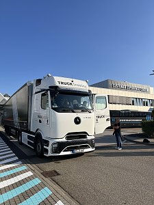 Ein weißer Mercedes-Benz Lkw mit Sattelauflieger steht bei strahlendem Sonnenschein vor einem Mercedes-Benz Gebäude. Die Fahrertür der Zugmaschine ist geöffnet, daneben steht eine Person.