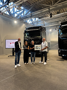 Vier Personen stehen nebeneinander für ein Übergabefoto vor einem großen, schwarzen Mercedes-Benz Lkw in einer hellen Werkshalle. Sie halten ein gerahmtes Bild sowie ein großen Schlüssel in der Hand.