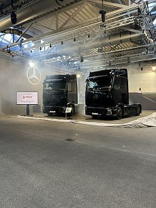 Zwei baugleiche, glänzende schwarze Mercedes-Benz Lkw stehen nebeneinander in einer hell erleuchteten Übergabehalle. An der linken Wand wird ein leuchtender Mercedes-Stern projiziert. Daneben steht ein Bildschirm mit dem Logo von Ehrlich Logistics.