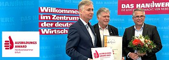Drei Männer stehen vor einer blauen Wand mit dem Schriftzug „Das Handwerk  Die Wirtschaftsmacht von nebenan“. Einer hält eine Urkunde, ein anderer eine Blumenstrauß, der dritte das Schild „Ausbildungsaward 2025 Handwerkskammer Erfurt“. Das Bild zeigt die Übergabe des Ausbildungsawards an die Autohaus Peter Gruppe. (Autohaus Peter)